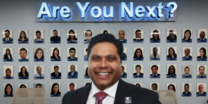 Dr. Michael Everest in a suit and tie standing in front of his “Are You Next?” wall in his conference room.